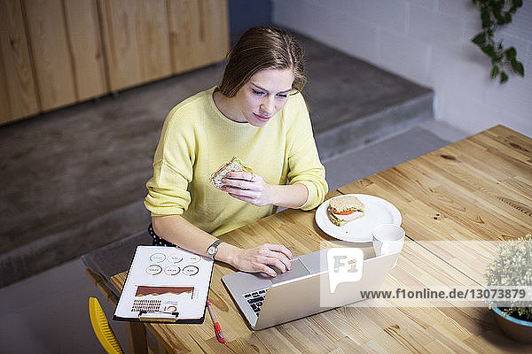 Hochwinkelansicht einer Geschäftsfrau  die ein Sandwich isst  während sie am Tisch einen Laptop benutzt
