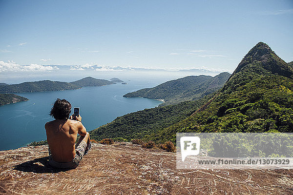 Rear view of man photographing sea while sitting on mountain against sky
