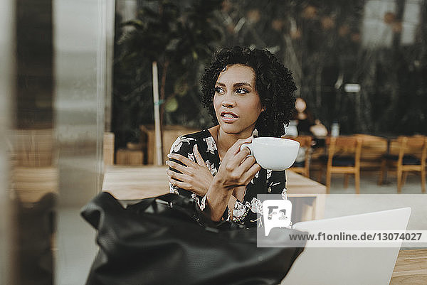 Nachdenkliche Frau hält Kaffeetasse in der Hand  während sie im Café am Laptop-Computer sitzt
