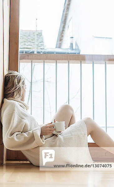 Seitenansicht einer Frau mit Kaffeetasse  die durch das Fenster schaut  während sie zu Hause auf dem Boden sitzt