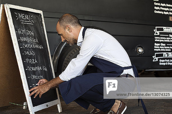 Side view of male vendor writing on blackboard while crouching at street by food truck
