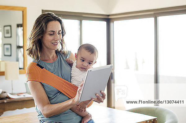 Mutter  die einen kleinen Jungen in einer Babytrage trägt  während sie zu Hause einen Tablet-Computer benutzt