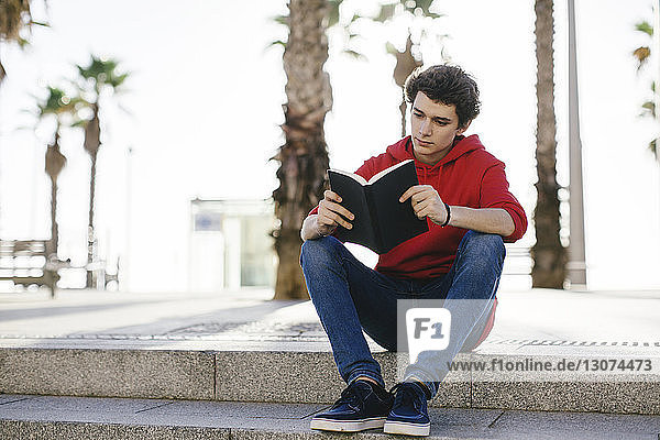 Teenage boy reading book while sitting on steps in city