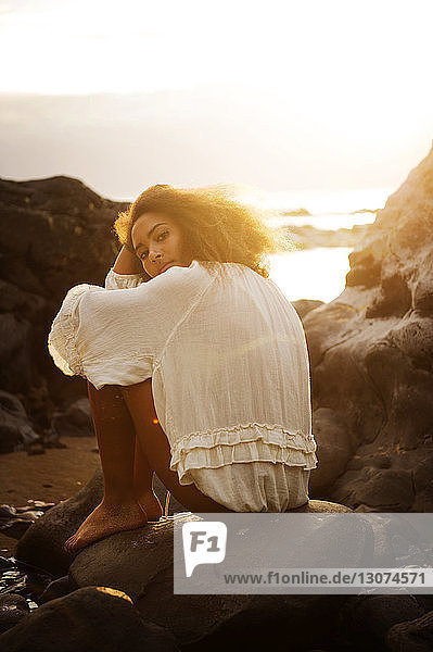 Portrait of teenage girl resting on rock at seashore during sunny day