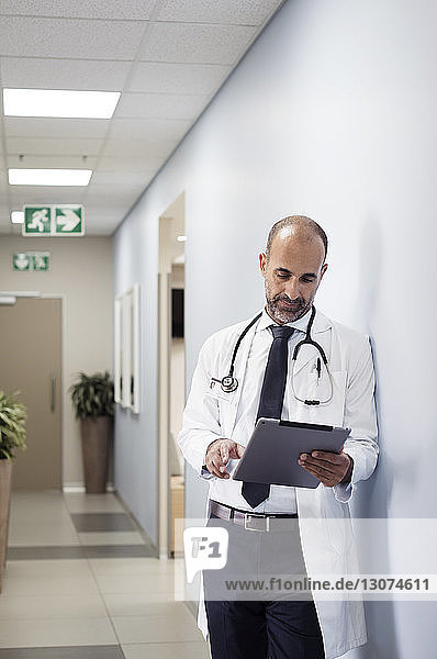 Doctor using tablet computer while leaning on wall in corridor