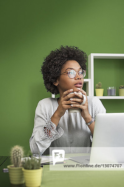 Nachdenkliche Frau mit Laptop-Computer beim Kaffee trinken im Büro
