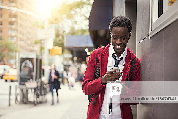 Smiling student using phone while leaning on wall in city