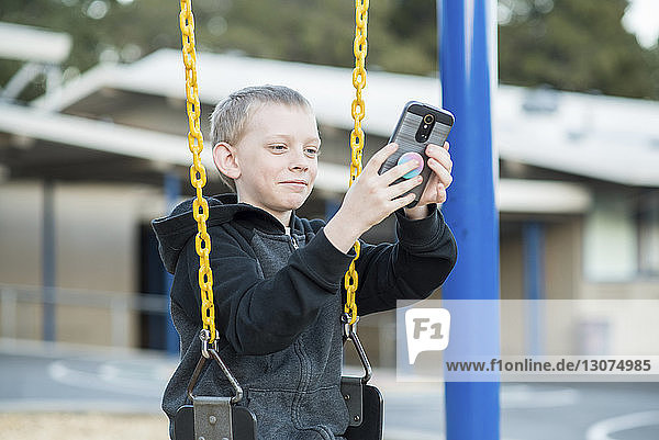 Niedrigwinkelansicht eines lächelnden Jungen  der ein Mobiltelefon benutzt  während er im Park schaukelt