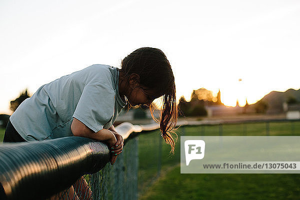 Seitenansicht eines Mädchens  das sich bei Sonnenuntergang auf dem Spielplatz über den Zaun beugt