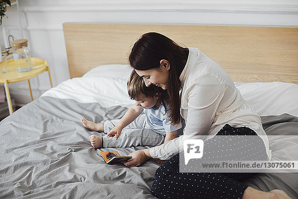Happy mother showing picture book to baby boy while sitting on bed at home