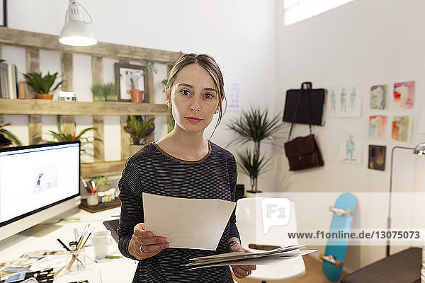 Portrait of confident female illustrator holding papers while standing in creative office