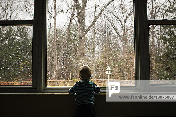 Rückansicht eines Kleinkindes  das im Winter von zu Hause durch ein Fenster schaut