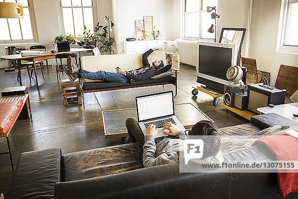 Hochwinkelansicht eines Geschäftsmannes mit Laptop  während ein Mitarbeiter im Kreativbüro auf dem Sofa liegt