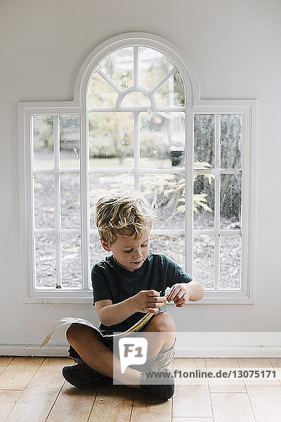 Junge mit Buch sitzt zu Hause auf dem Boden gegen das Fenster