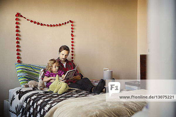 Vater zeigt einem Mädchen ein Buch  während er zu Hause auf dem Bett sitzt