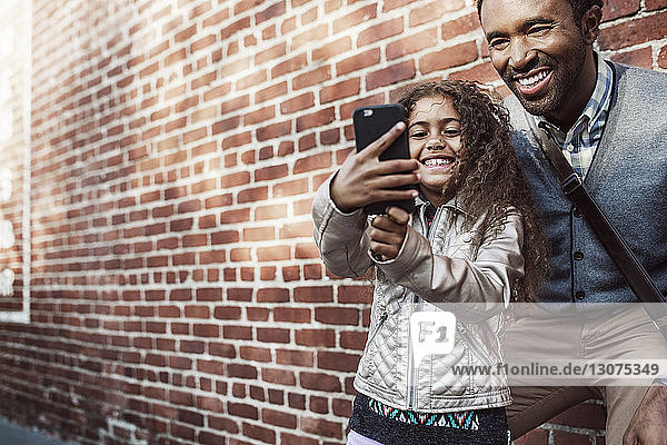 Daughter taking selfie with father through smart phone against brick wall