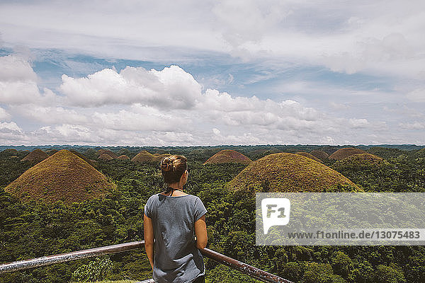 Rear view of woman looking at Chocolate Hills while standing by railing against cloudy sky