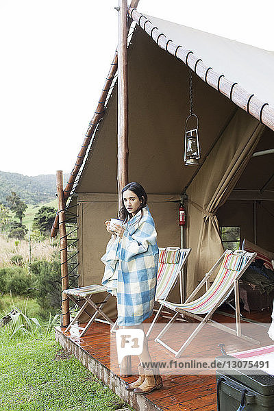 In ein Handtuch gewickelte Frau trinkt Kaffee  während sie während des Urlaubs im Zelt steht