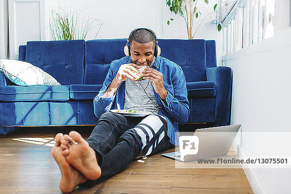 Man eating sandwich while listening music through laptop computer by sofa at home
