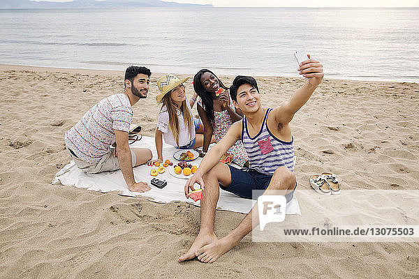 Man taking selfie with friends while enjoying picnic on beach