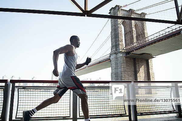 Side view of young man jogging by railing against Brooklyn Bridge