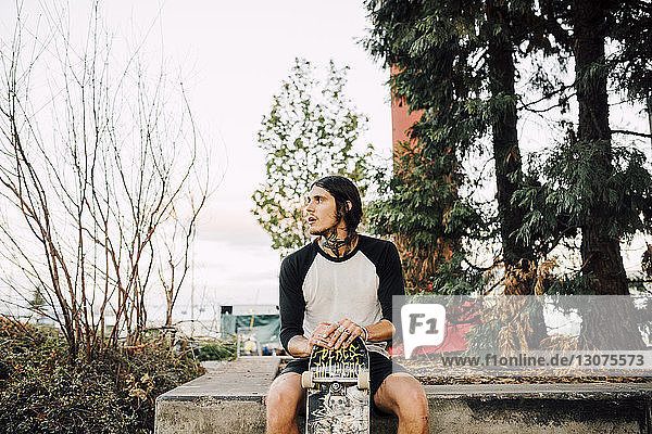 Man with skateboard looking away while sitting on retaining wall against sky in city