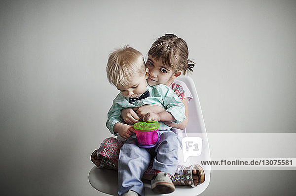 Porträt eines niedlichen Mädchens  das mit seinem Bruder auf einem Stuhl vor einer weißen Wand zu Hause sitzt