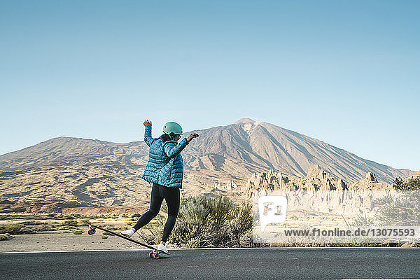 Rückansicht einer Frau  die einen Stunt auf dem Skateboard gegen Berge vorführt