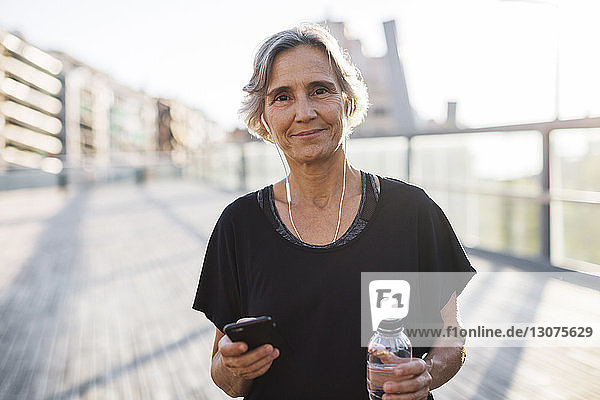 Portrait of confident woman holding smart phone and water bottle at bridge