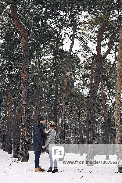 Happy couple looking at each other while standing on snow covered field in forest