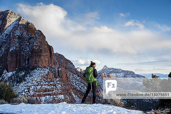 Rückansicht eines Mannes mit grünem Rucksack  der auf Schnee in der Wüste vor Felsformationen und bewölktem Himmel im Winter steht