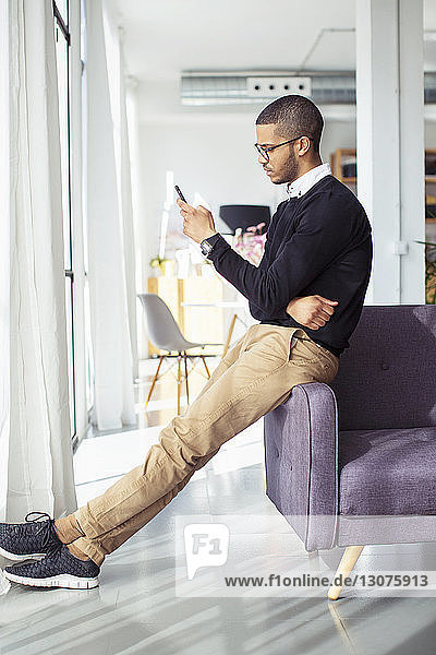 Seitenansicht eines Geschäftsmannes  der ein Smartphone benutzt  während er sich im Büro auf ein Sofa lehnt