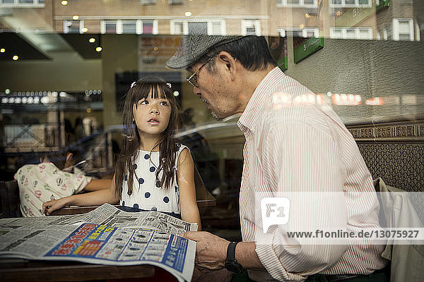 Girl looking at grandfather while sitting in restaurant seen through glass