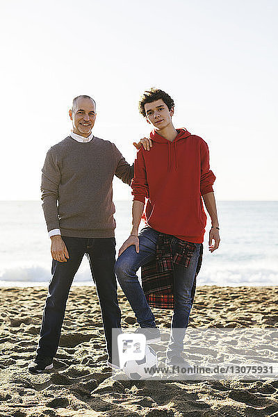 Porträt eines selbstbewussten Sohnes und Vaters mit Fussball am Strand vor klarem Himmel