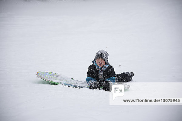 Sorgloser Junge liegt bei Schneefall mit Schlitten auf verschneitem Feld