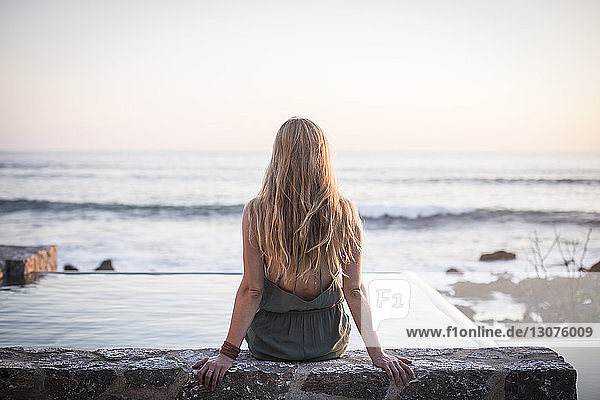 Rückansicht einer Frau  die auf einer Stützmauer an einem künstlichen Teich gegen das Meer und den klaren Himmel sitzt
