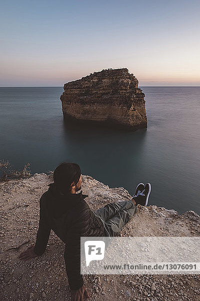 High angle view of man looking at sea while sitting on rock formation against clear sky during sunset