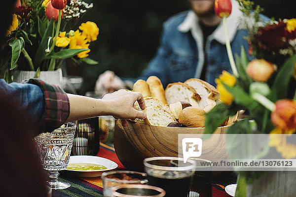 Cropped image of woman taking bread from wooden bowl while sitting with friend at table