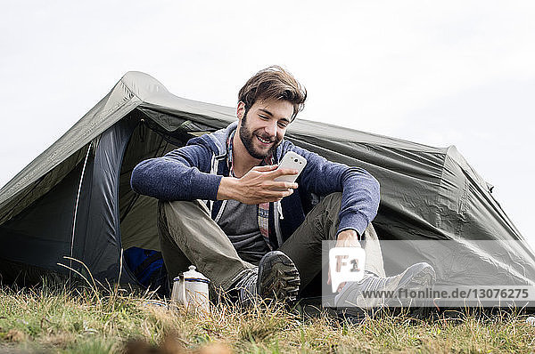 Smiling man using smart phone outside tent on hill against clear sky