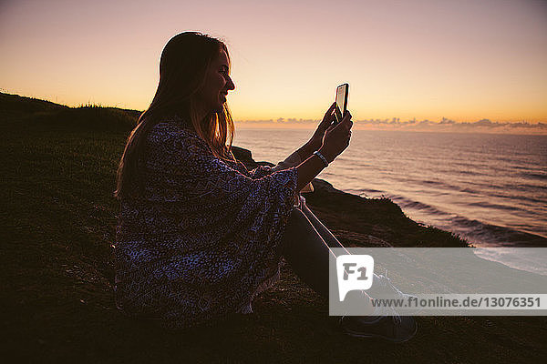 Side view of woman photographing through smart phone while sitting on hill by sea during sunset