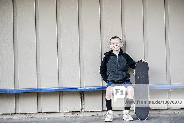 Portrait of smiling boy holding skateboard while sitting on bench against wall