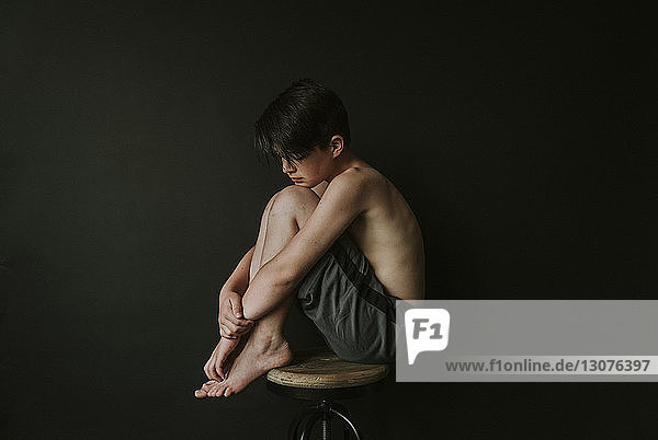 Seitenansicht eines traurigen Jungen ohne Hemd  der auf einem Hocker vor schwarzem Hintergrund sitzt