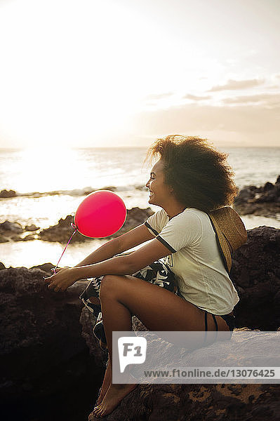 Happy teenage girl holding balloon while sitting on rocky shore