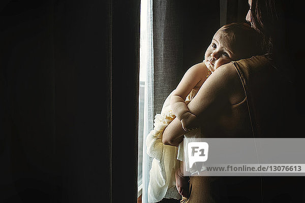Mother carrying daughter while standing by window at home