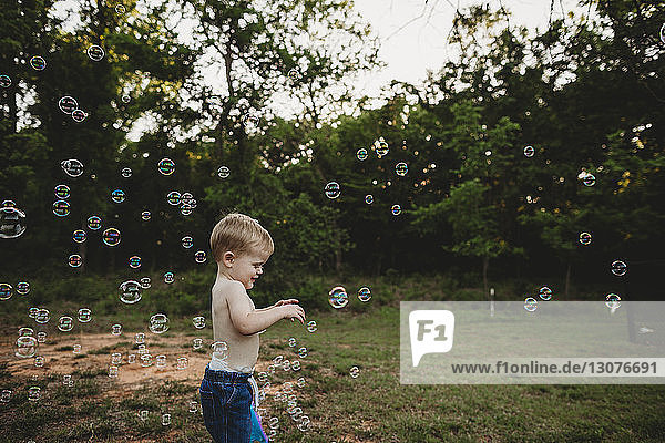 Side view of shirtless baby boy playing with bubbles while standing at park