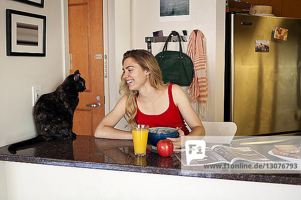 Lächelnde Frau sieht Katze an  während sie beim Frühstück auf der Küchentheke sitzt