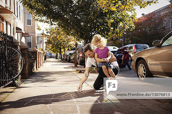 Father carrying daughter while drawing hopscotch on footpath