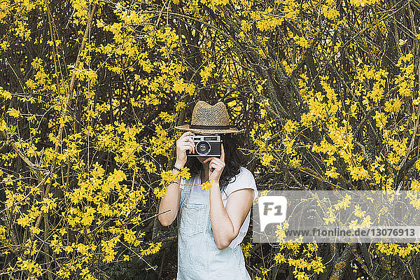 Junge Frau fotografiert mit einer Oldtimer-Kamera  während sie inmitten von gelben Blüten auf Zweigen im Park steht