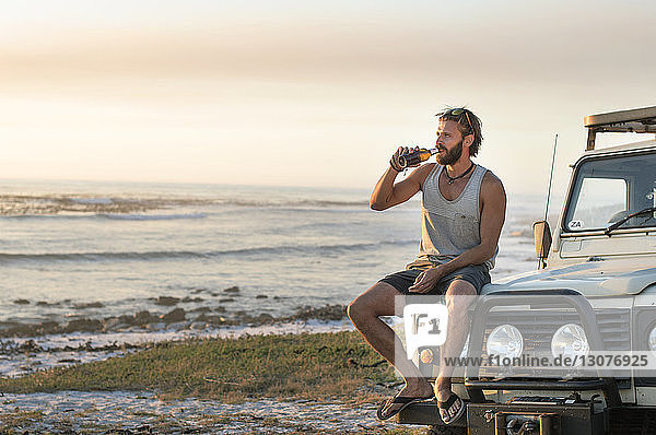 Mann trinkt Bier  während er bei Sonnenuntergang auf einem Geländewagen am Strand sitzt