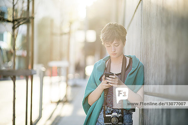 Teenage girl using smart phone while leaning on wall in city